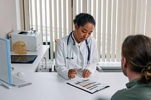 A doctor sitting with patient in an office in front of window.