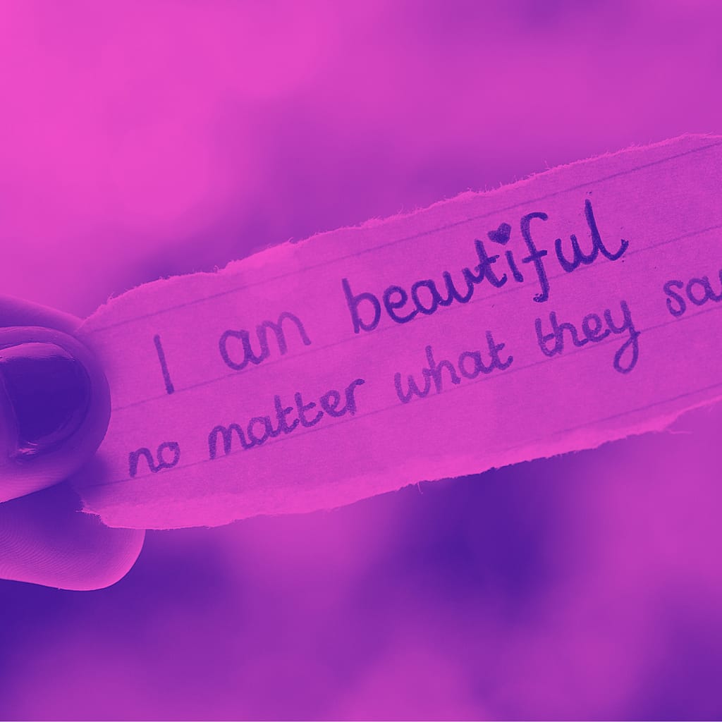 Image of fingers holding a ripped piece of paper that says I am beautiful no matter what they say.